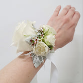 A wrist corsage featuring a white floral arrangement with green leaves and red accents, attached to a white ribbon.