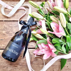 A wrapped bottle of Bird in Hand sparkling wine placed next to a bouquet of pink lilies on a wooden surface.