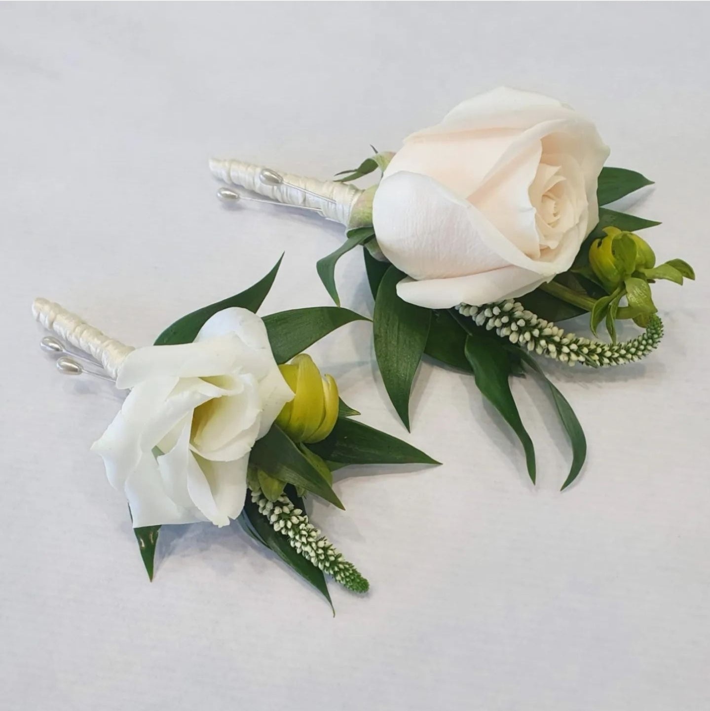 A floral buttonhole featuring a white rose, green leaves, and white ribbon.