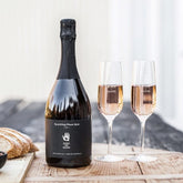 A bottle of Bird In Hand Sparkling Wine with two filled champagne glasses in the background, placed on a wooden surface with a neutral-colored backdrop.