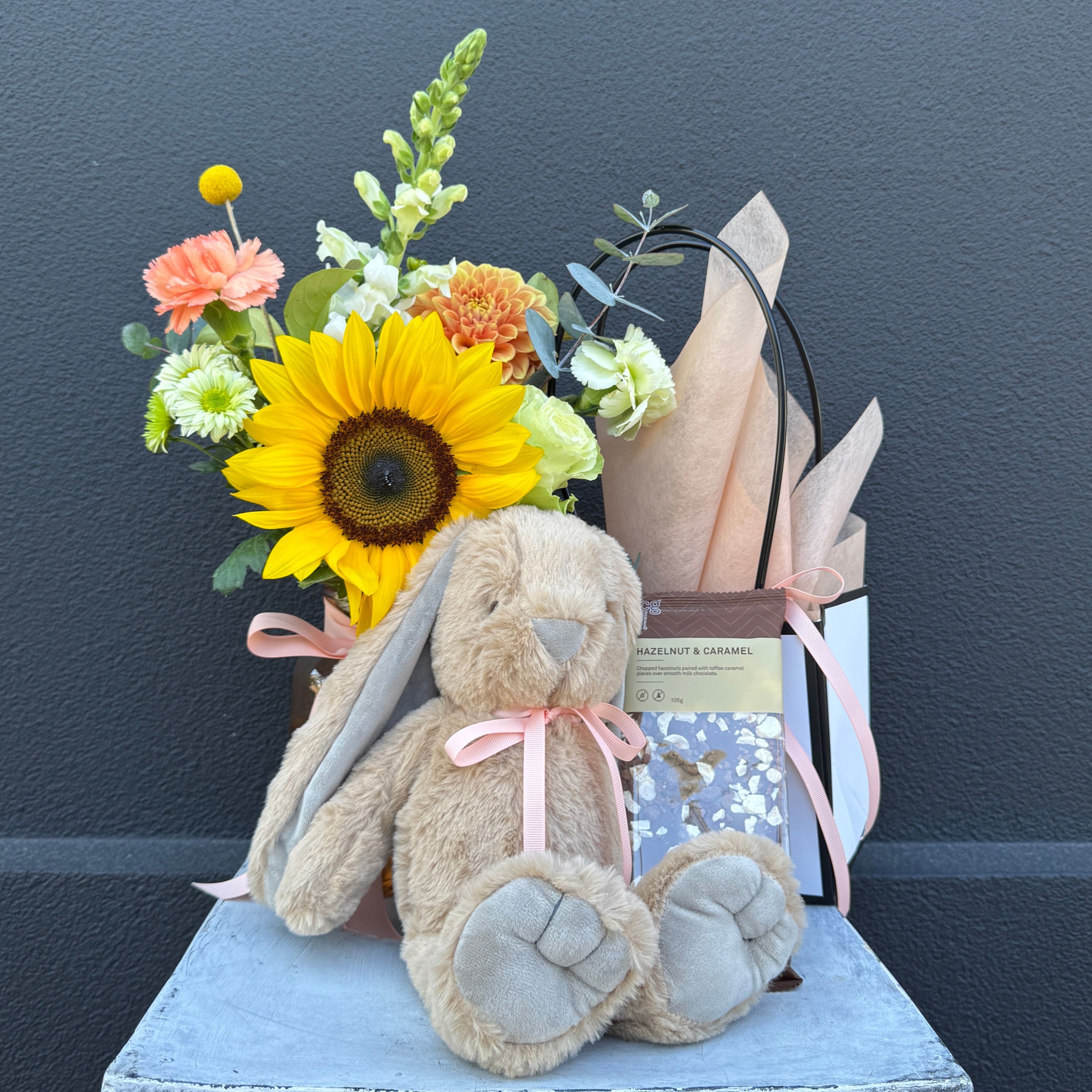 A gift bag setup with a plush bunny, a posy of flowers including a sunflower, and some packaging elements like a ribbon and card, against a dark background.