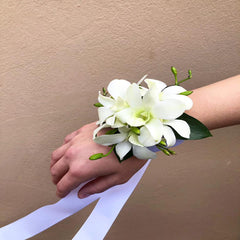 A wrist corsage featuring a white floral arrangement with green leaves and red accents, attached to a white ribbon.
