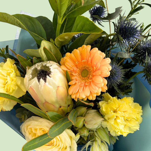 Bouquet of flowers including orange gerbera, yellow carnations, and protea on a light green background.