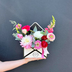 A hand holding a fresh flower arrangement in the shape of an envelope, with a variety of colors including pink, red, white, and green.
